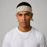 A man wearing a beige patterned headband with leaf motifs, a white t-shirt, and a silver chain necklace, photographed against a neutral gray background.