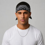 A man wearing a black bandana with a white paisley pattern tied around his head, paired with a white t-shirt and a silver chain necklace, photographed against a neutral gray background.