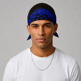 A man wearing a blue and black paisley patterned headband with matching blue ear cuffs, paired with a white t-shirt and a silver chain necklace, set against a plain gray background.