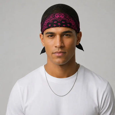A man wearing a black bandana with a pink paisley pattern and a black baseball cap, paired with a white t-shirt and a silver chain necklace, set against a plain light gray background.
