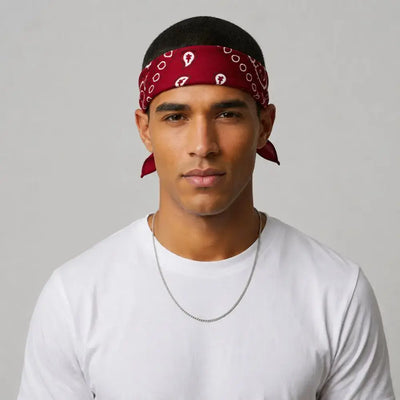 A man wearing a red blood bandana with a white paisley pattern tied around his head, paired with a white t-shirt and a silver chain necklace, set against a plain light gray background.