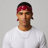 A man wearing a red blood bandana with a white paisley pattern tied around his head, paired with a white t-shirt and a silver chain necklace, set against a plain light gray background.
