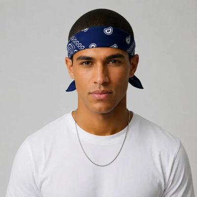 A man wearing a navy blue bandana with white paisley patterns tied around his head, paired with a white t-shirt and a silver chain necklace, set against a plain light gray background.