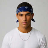 A man wearing a navy blue bandana with white paisley patterns tied around his head, paired with a white t-shirt and a silver chain necklace, set against a plain light gray background.