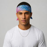 A man wearing a blue and pink paisley bandana headwrap with a white t-shirt and a silver chain necklace, photographed against a neutral light gray background.