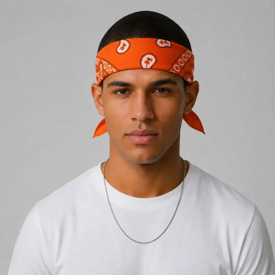 A man wearing a bright orange paisley bandana headwrap with matching orange ear cuffs, paired with a white t-shirt and a thin silver chain necklace, set against a plain light gray background.