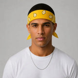 A man wearing a yellow bandana headband with a paisley pattern and matching yellow ear loops, paired with a white t-shirt and a silver chain necklace, set against a plain light gray background.