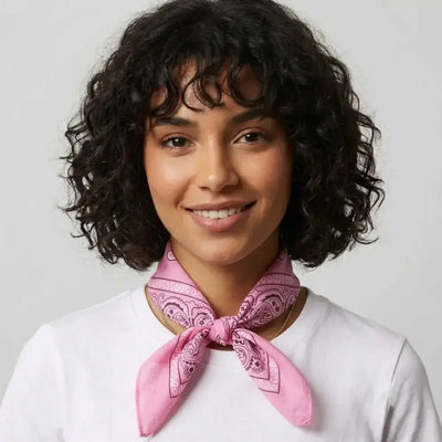 A woman with short curly dark hair wearing a white t-shirt and a pink paisley bandana tied around her neck, set against a plain light gray background.