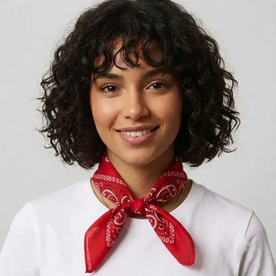 A woman with short curly dark hair wearing a red bandana tied around her neck and a white t-shirt, photographed against a light gray background.