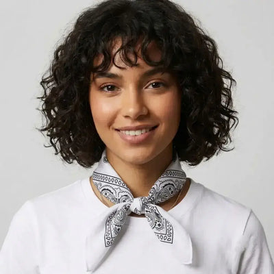 A woman with short curly dark hair wearing a white crew-neck top and a black and white paisley bandana tied around her neck, set against a plain light gray background.