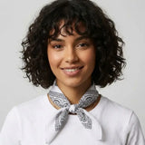A woman with short curly dark hair wearing a white crew-neck top and a black and white paisley bandana tied around her neck, set against a plain light gray background.